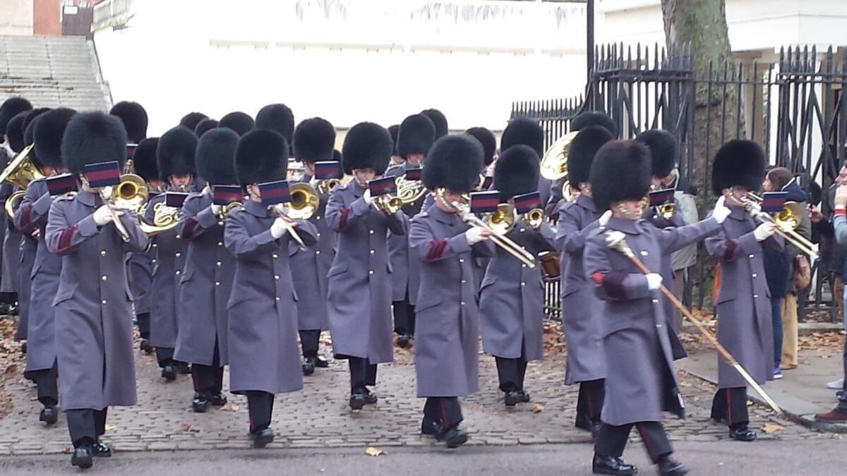 The Band of the Household Division marching in grey winter greatcoats past iron railings