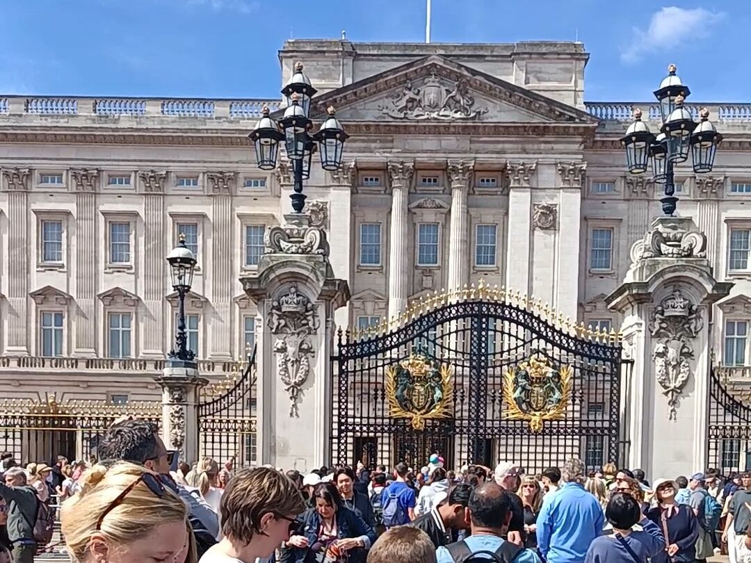 Crowds packed against the gates of Buckingham Palace beneath the gold royal crests, waiting for Changing the Guard
